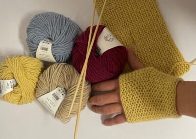 4 balls of yarn in different shades, a hand wearing a knitted handwarmer, knitting needles and a knitted rectangle